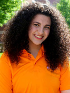 A smiling young woman with dramatically curly long dark brown hair wearing a UT orange polo shirt