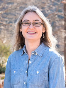 A smiling woman in glasses and a blue chambray shirt stands outside in the sun, surrounded by nature