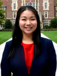 A smiling young Asian woman with long dark hair stands in front of Ayres Hall wearing a blue blazer over a red shirt