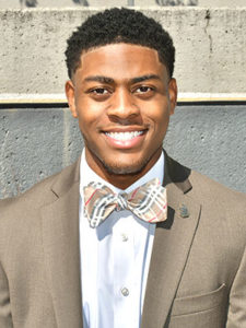 A smiling young Black man with short hair wearing a tan suit jacket and spiffy plaid bow tie