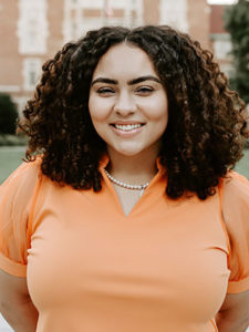 A smiling young woman with curly brown hair wearing an orange polo shirt