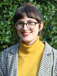 A smiling young woman with dark brown hair pulled back wearing funky glasses and a bright yellow turtleneck under a stylish houndstooth blazer