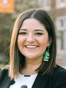A smiling young woman with straight brown hair wearing awesomely funky green earrings and a black blazer