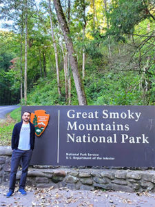 A man wearing a sweater standing beside the entrance sign to the Great Smoky Mountains National Park